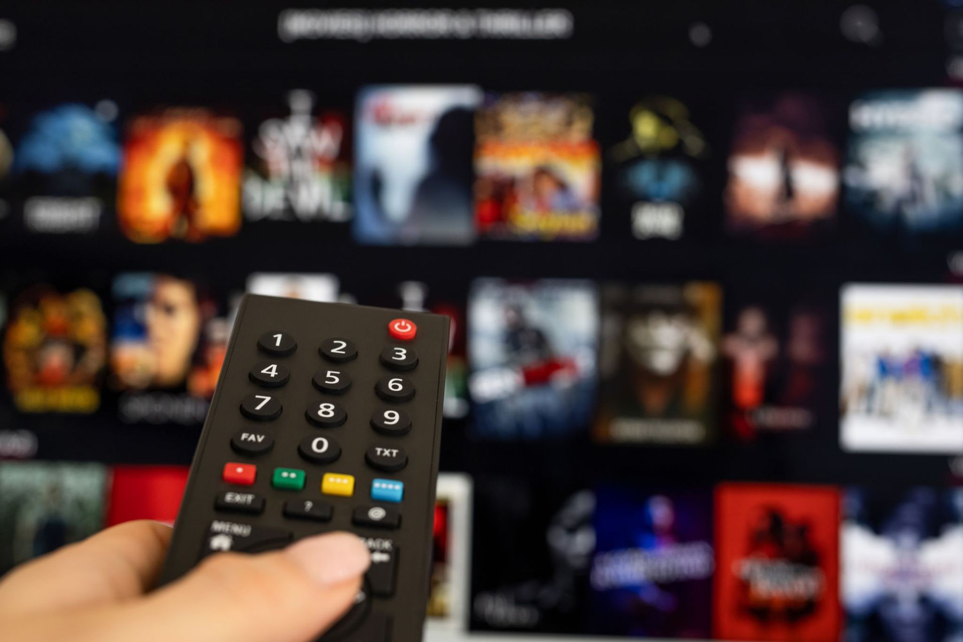 Hand holding a remote control in front of a TV screen displaying streaming service movie thumbnails.